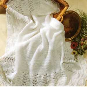 Peut inclure: Une couverture tricotée blanche présentant un motif ondulé délicat sur toute la partie centrale et une bordure en dentelle festonnée décorative sur les bords. Le tissu a une texture douce avec des mailles visibles, créant un aspect léger.