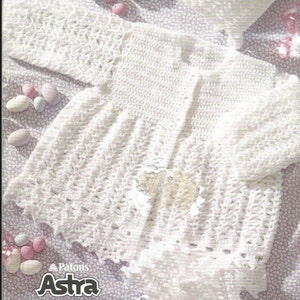 May include: A white crocheted long-sleeve cardigan with a textured pattern, a round neckline, and a button-front closure. The garment is displayed alongside a matching crocheted bonnet and booties. Text on the image reads Patons Astra and Patons Fairytale.