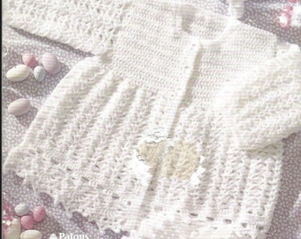 May include: A white crocheted long-sleeve cardigan with a textured pattern, a round neckline, and a button-front closure. The garment is displayed alongside a matching crocheted bonnet and booties. Text on the image reads Patons Astra and Patons Fairytale.
