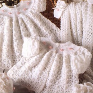 May include: A set of white knitted garments including two dresses, a bonnet, and a blanket, all featuring an intricate openwork pattern. The dresses have short sleeves and are decorated with small, pale pink ribbon roses along the neckline.