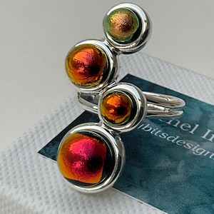 May include: A silver ring featuring four circular, iridescent glass cabochons arranged in a vertical, staggered line. The cabochons display shifting colours of red, orange, and green. The ring band is made of polished silver metal.
