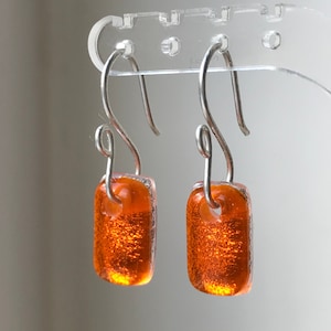 Peut inclure: Une paire de boucles d'oreilles rectangulaires orange avec des crochets argentés. Les boucles d'oreilles ont un aspect texturé et scintillant, et les crochets sont incurvés. Les boucles d'oreilles sont exposées sur un présentoir transparent.