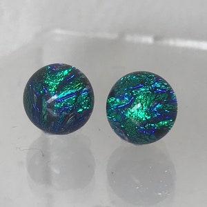 Può includere: Un paio di orecchini rotondi in vetro iridescente con uno schema di colori blu e verde. Gli orecchini hanno un aspetto testurizzato e marmorizzato.
