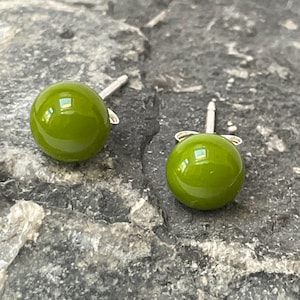 May include: A pair of green glass ball stud earrings with silver posts. The earrings are on a grey textured surface.