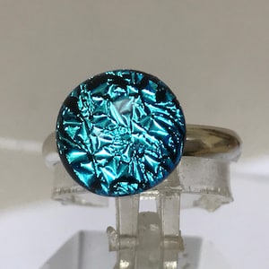 Pode incluir: Um anel de vidro dicróico azul turquesa com uma banda prateada. O vidro tem um acabamento texturizado e iridescente.