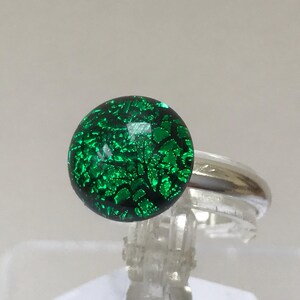 Pode incluir: Um anel de prata com um cabochão de vidro verde. O vidro tem uma base verde escura com um padrão de manchas verdes mais claras e prateadas.