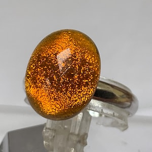 Pode incluir: Um anel de vidro dicróico com uma banda prateada. O anel apresenta uma pedra grande, oval, de vidro dicróico laranja e dourado. O vidro tem uma aparência texturizada e salpicada, refletindo a luz. O anel está sobre um expositor transparente.