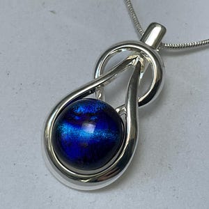 May include: A silver pendant necklace with a teardrop design and a striking blue orb. The pendant hangs from a fine silver chain. The orb has an iridescent sheen, displaying hues of blue and purple. The necklace is a stylish accessory.