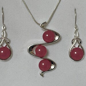 May include: A silver necklace and earrings set with three pink gemstone beads. The necklace features a wavy design with the beads set in a row.