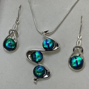 以下が含まれることがあります： ネックレス、イヤリング、ペンダントのシルバーのジュエリーセット。セットには、3つの虹色の青と緑のオーブが付いたペンダント付きネックレスが含まれています。イヤリングとペンダントは同様のデザインで、それぞれに1つのオーブが付いています。