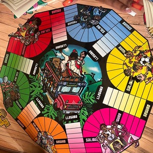 May include: A colourful game board with a red, yellow, green, and pink track. The board features illustrations of people, a bus, and a jungle scene. The words "LLEGAGA", "SALIDA", and "SEGURO" are printed on the board.