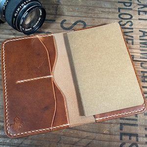May include: Open, brown leather wallet with a notebook insert. The wallet has card slots and is stitched with white thread. A camera lens is visible in the upper left corner.