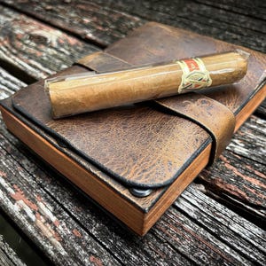 May include: A brown leather journal with a wooden cover and a cigar inside. The journal has a strap closure and a distressed leather look.