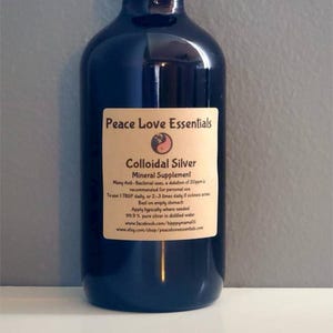 Op de afbeelding: Een donkerblauwe glazen fles met een zwart etiket waarop "Peace Love Essentials Colloïdaal Zilver Mineraal Supplement" staat. Het etiket bevat ook gebruiksaanwijzingen en contactgegevens.