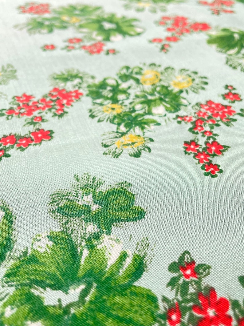 May include: A light blue fabric with a repeating pattern of green leaves and red flowers. The fabric has a subtle texture.