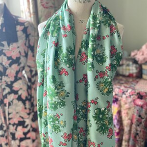 May include: A light green fabric with a floral print featuring green leaves, white flowers, and red berries. The fabric is draped over a dress form.