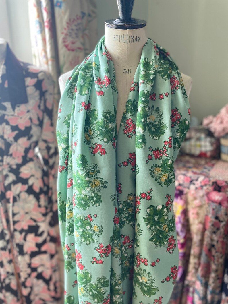May include: A light green fabric with a floral print featuring green leaves, white flowers, and red berries. The fabric is draped over a dress form.