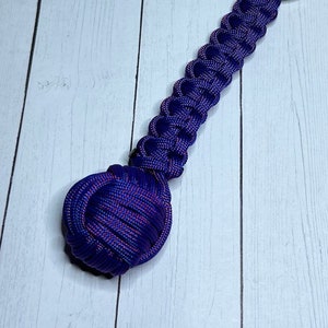 May include: A purple and pink paracord keychain with a knotted ball at the end. The keychain has a silver key ring.