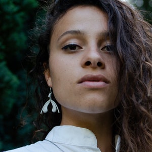 May include: A woman with long, curly brown hair wearing a white collared shirt with a silver chain necklace. She is wearing silver earrings shaped like a pair of hands.