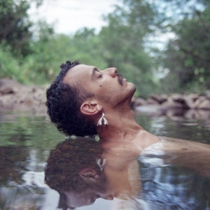May include: A person with dark hair floats face up in a shallow stream of water. The person is wearing a pair of small white earrings.