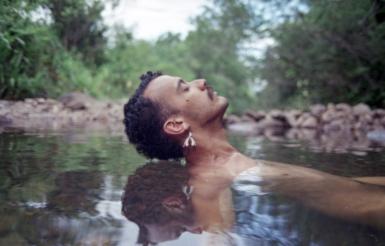 May include: A person with dark hair floats face up in a shallow stream of water. The person is wearing a pair of small white earrings.