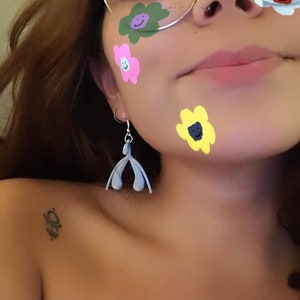 May include: A pair of blue dangle earrings shaped like a vulva. The earrings are hanging from the earlobe of a person with brown hair. The person has a few colorful flower stickers on their face.