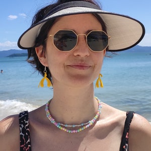 May include: A woman wearing a black visor, sunglasses, and a multi-colored beaded necklace. She is wearing yellow earrings shaped like a stylized flower.