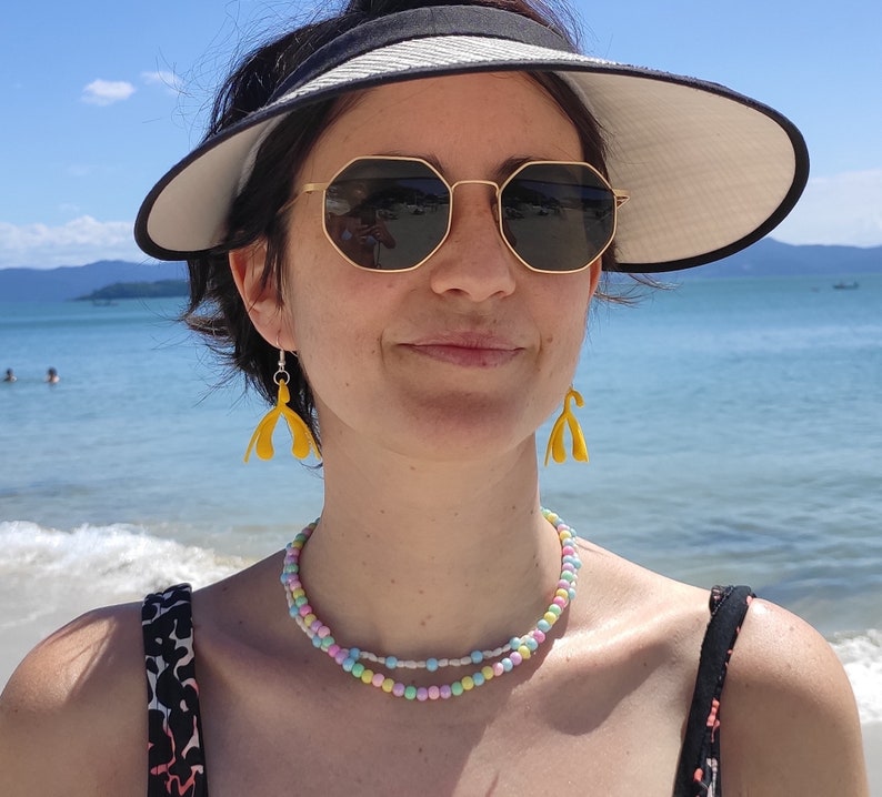 May include: A woman wearing a black visor, sunglasses, and a multi-colored beaded necklace. She is wearing yellow earrings shaped like a stylized flower.