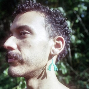 May include: A man with curly dark hair and a beard is wearing a green plastic earring shaped like a hand. He is standing in front of a blurred green background.