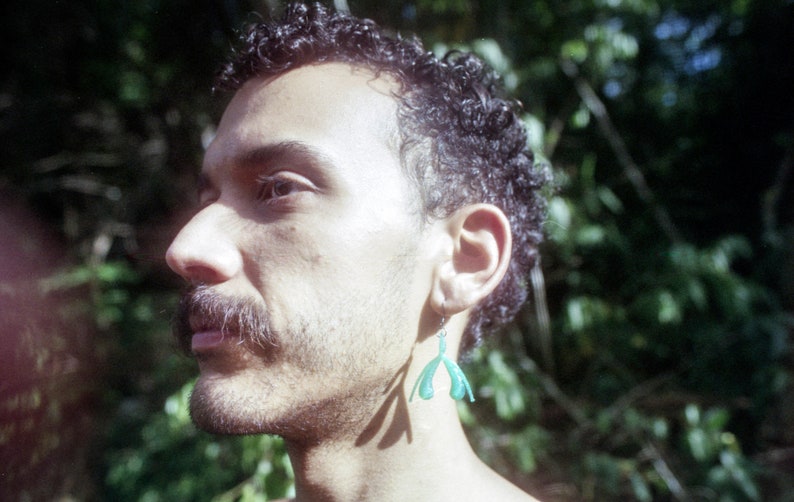 May include: A man with curly dark hair and a beard is wearing a green plastic earring shaped like a hand. He is standing in front of a blurred green background.