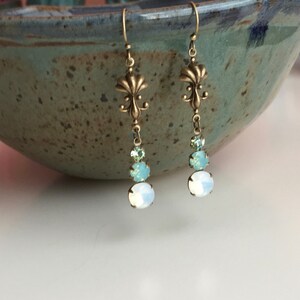 May include: A pair of dangle earrings with a fleur-de-lis charm and three crystal beads in shades of light blue and white.