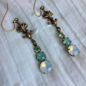 May include: A pair of dangle earrings with a gold fleur-de-lis charm and three iridescent stones. The stones are a light green, a darker green, and a white opal.