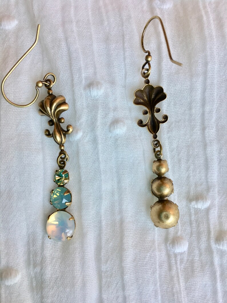May include: A pair of gold tone dangle earrings with a floral design and three round iridescent stones. The stones are a light green, a light blue, and a white opal.