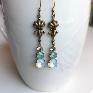 May include: A pair of dangle earrings with a bronze fleur-de-lis design and three crystal drops in shades of green and white.