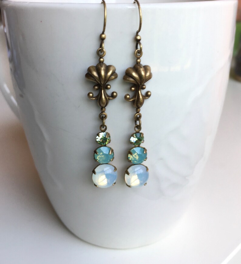 May include: A pair of dangle earrings with a bronze fleur-de-lis design and three crystal drops in shades of green and white.