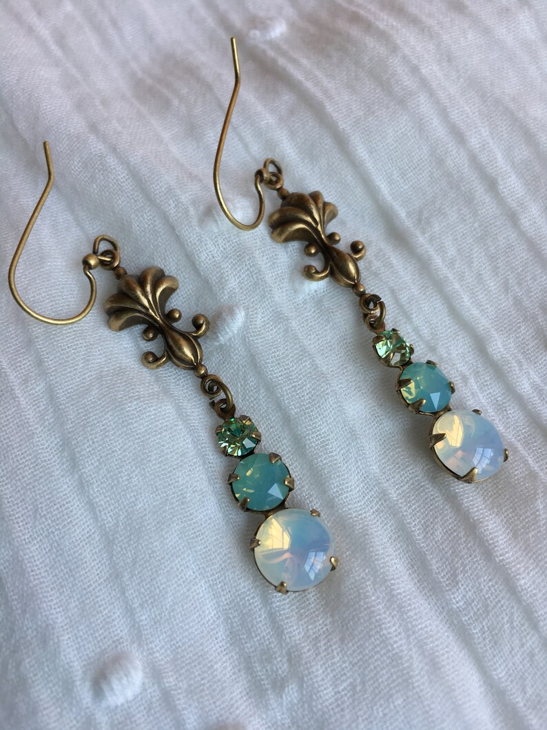 May include: A pair of dangle earrings with a gold fleur-de-lis charm and three iridescent stones. The stones are a light green, a darker green, and a white opal.