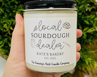 May include: A white candle in a glass jar with a black lid. The label reads "local sourdough dealer" with illustrations. The candle is held in front of a woven basket and greenery. The text "Kate's Bakery Est. 2023" is also on the label.