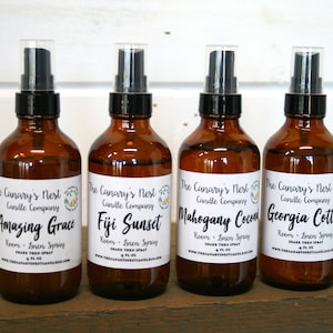 May include: Four amber glass bottles with black spray tops and white labels. The labels have the text "The Canary's Nest Candle Company" and the names of the scents: "Amazing Grace", "Fiji Sunset", "Mahogany Coconut", and "Georgia Cotton". Each label also says "Room - Linen Spray" and "4 fl oz".