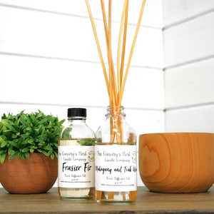 May include: Two glass bottles of reed diffuser oil with black lids and white labels. The labels read "The Canary's Nest Candle Company" and "Frasier Fir Reed Diffuser Oil" and "Mahogany and Teak Wood Reed Diffuser Oil". The bottles are sitting on a wooden surface with a small potted plant and a wooden bowl.