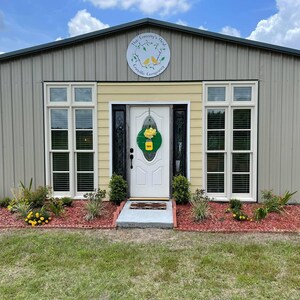 May include: A small, gray metal building with a yellow accent above the door. The building has two windows on each side of the door and a sign above the door that reads "The Country Nest Candle Company".