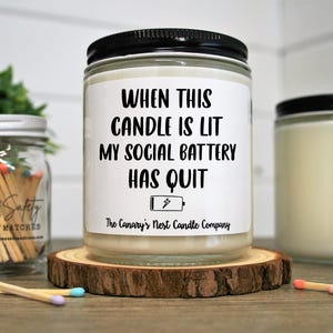 Peut inclure: Une bougie en pot de verre avec un couvercle noir et une &eacute;tiquette blanche indiquant "WHEN THIS CANDLE IS LIT MY SOCIAL BATTERY HAS QUIT". La bougie repose sur un dessous de verre en bois. Un pot d'allumettes est en arri&egrave;re-plan.