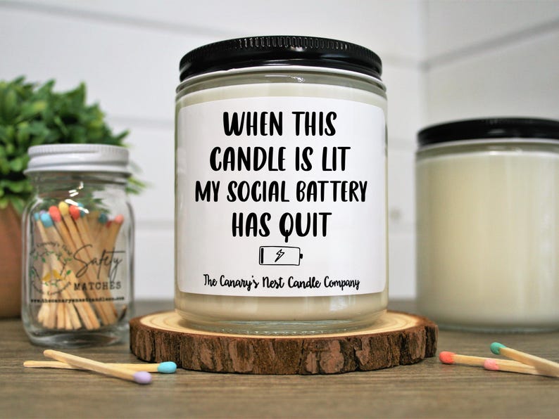 Peut inclure: Une bougie en pot de verre avec un couvercle noir et une &eacute;tiquette blanche indiquant "WHEN THIS CANDLE IS LIT MY SOCIAL BATTERY HAS QUIT". La bougie repose sur un dessous de verre en bois. Un pot d'allumettes est en arri&egrave;re-plan.