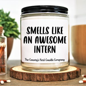 May include: A white candle with a black lid and label that reads "Smells Like An Awesome Intern" and "The Canary's Nest Candle Company".