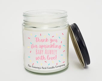 May include: A white candle in a clear glass jar with a black lid. The label on the jar says "Thank you for sprinkling Baby Aubrey with love!" and "The Canary's Nest Candle Company".