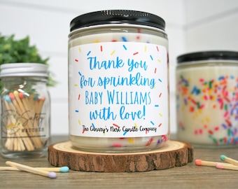 May include: Two glass jars with white candles decorated with colorful sprinkles. The jar on the left has a label that says "Thank you for sprinkling Baby Williams with love! The Canary's Nest Candle Company".