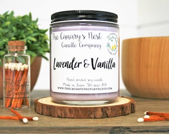 May include: A glass jar candle with a purple soy wax and a black lid. The label reads "The Canary's Nest Candle Company" and "Lavender & Vanilla". The candle is sitting on a wooden coaster.