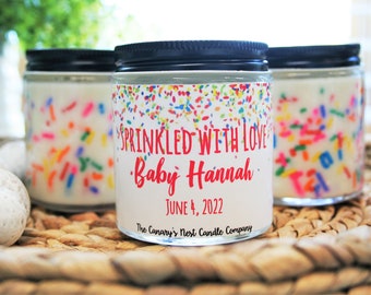 May include: Three glass candle jars with white labels and colorful sprinkles. The label on the center jar reads "SPRINKLED WITH LOVE Baby Hannah JUNE 4, 2022 The Canary's Nest Candle Company".
