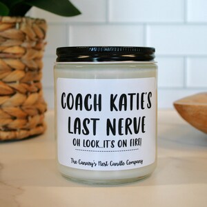 May include: A white candle with a black label that reads "Coach Katie's Last Nerve Oh look... it's on fire!" The Canary's Nest Candle Company.
