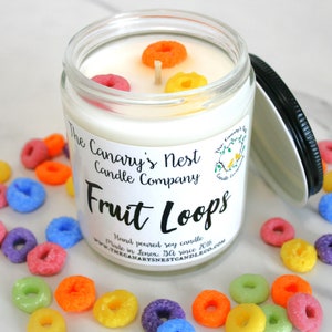 May include: A glass jar candle with a black lid, labeled "Fruit Loops" by The Canary's Nest Candle Company. The candle is white with colorful cereal pieces on top. Scattered around the jar are more colorful cereal pieces.