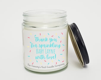 May include: A clear glass jar candle with a black lid, featuring a label with the text "Thank you for sprinkling BABY LAYNE with love!" in blue and yellow, surrounded by colorful confetti. The Canary's Nest Candle Company is printed at the bottom.
